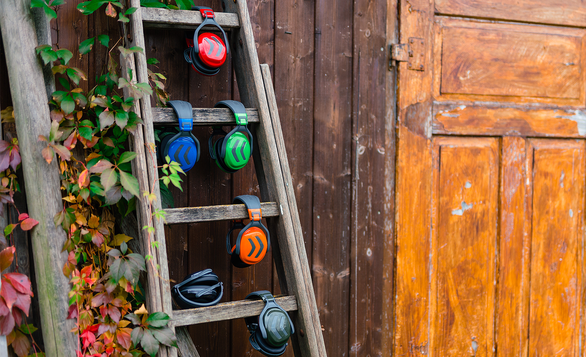 Six protections auditives colores suspendues  une vieille chelle en bois devant une faade en bois recouverte de feuillage automnal.