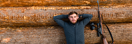 Un homme est allong dtendu, les bras croiss derrire la tte, sur des troncs d’arbres empils ;  ct de lui se trouve un fusil de chasse avec une lunette.