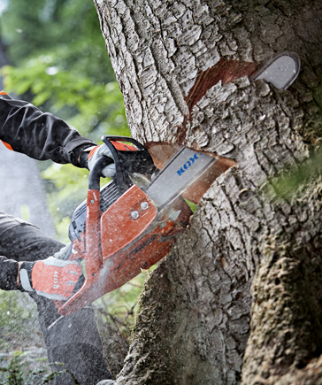 Une personne trononne un tronc d’arbre avec une trononneuse KOX, des copeaux de bois volent autour.