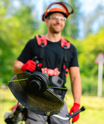 Homme avec casque et lunettes de protection tenant une dbroussailleuse  fil, sur fond de verdure.