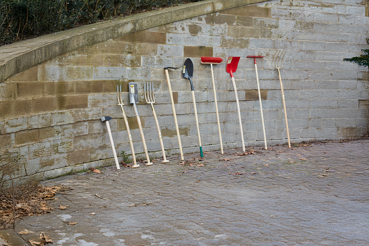 Plusieurs outils de jardinage � manche en bois sont appuy�s contre un mur en pierre, dont un marteau, des fourches-b�ches, une pelle, un balai, une pelle � neige, un r�teau et une fourche.