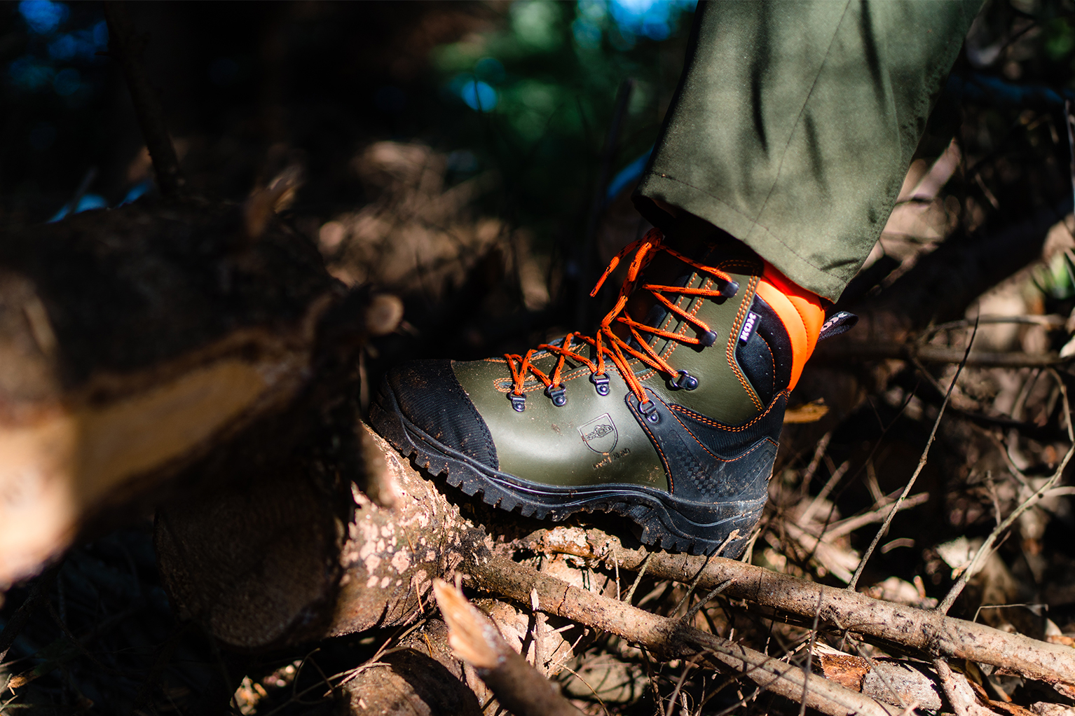 Une chaussure foresti�re robuste avec des lacets orange repose sur des branches et du bois en for�t, avec une semelle crant�e et une tige montante bien visibles.