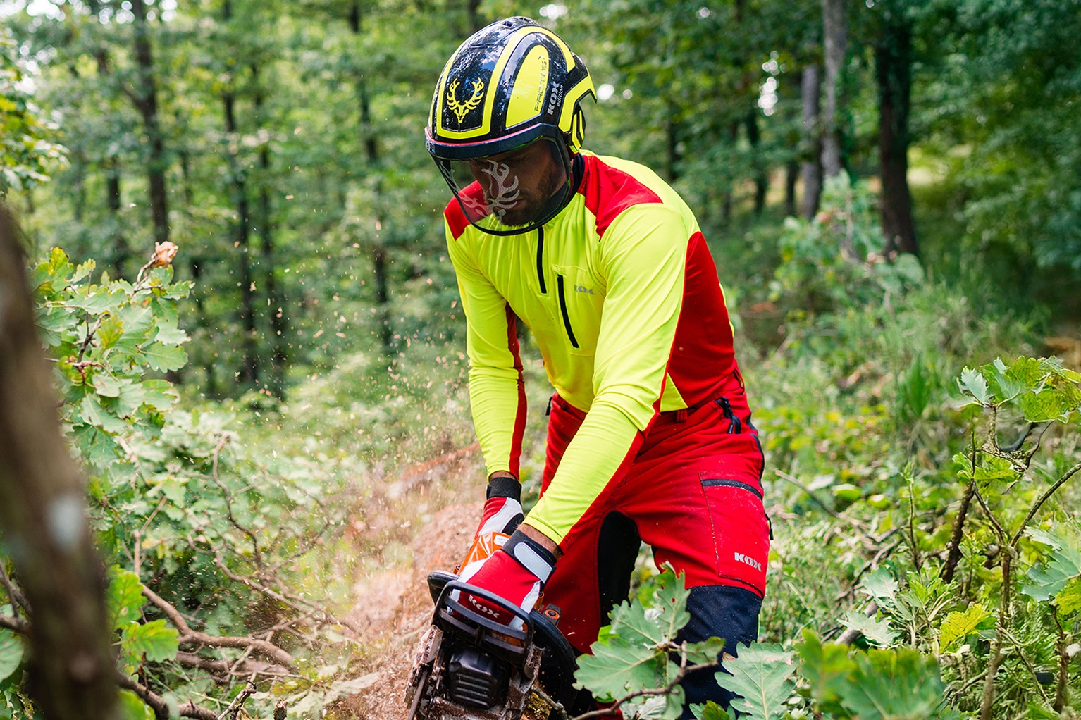 Travailleur forestier en pleine action avec une trononneuse, portant un t-shirt technique KOX orange vif avec des empicements noirs et un casque de protection avec visire et protections auditives.