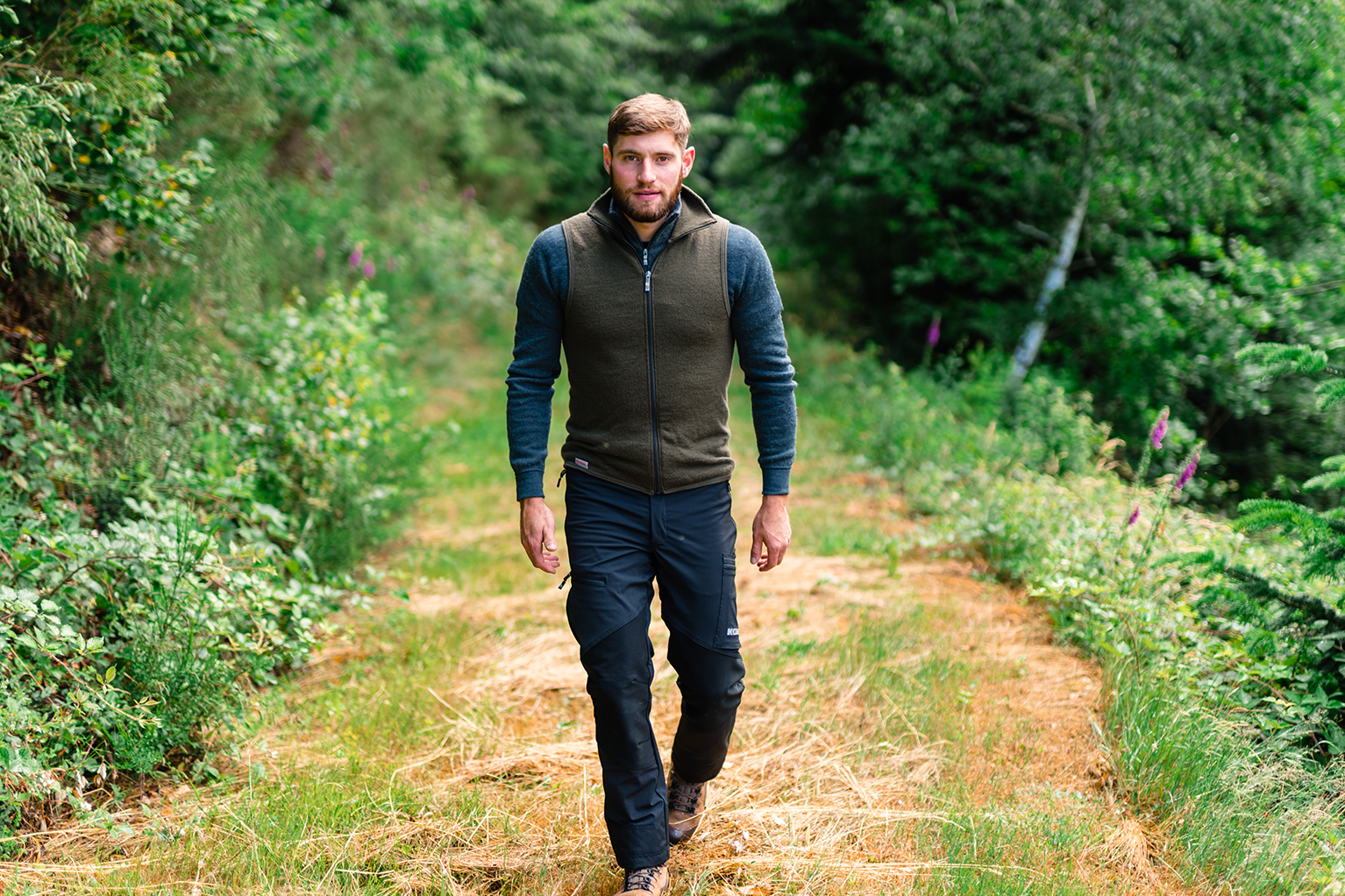 Un homme marche sur un sentier forestier, entour d’une fort verdoyante. Il porte une veste en polaire, un pantalon de plein air et des chaussures de randonne.