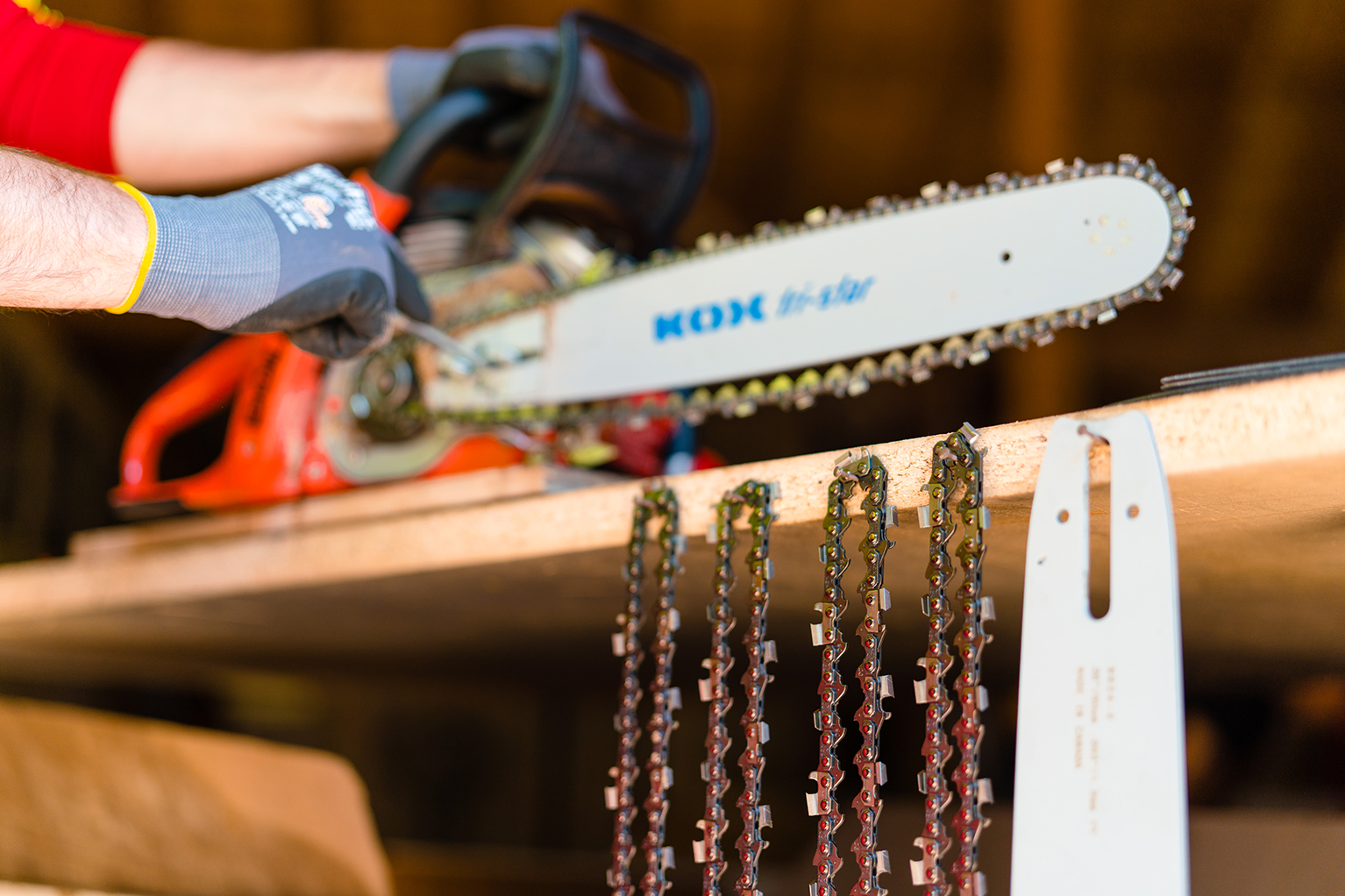 Personne portant des gants en train de tendre une chane de trononneuse KOX sur une trononneuse, au premier plan plusieurs chanes de rechange et un guide suspendus  une tagre.