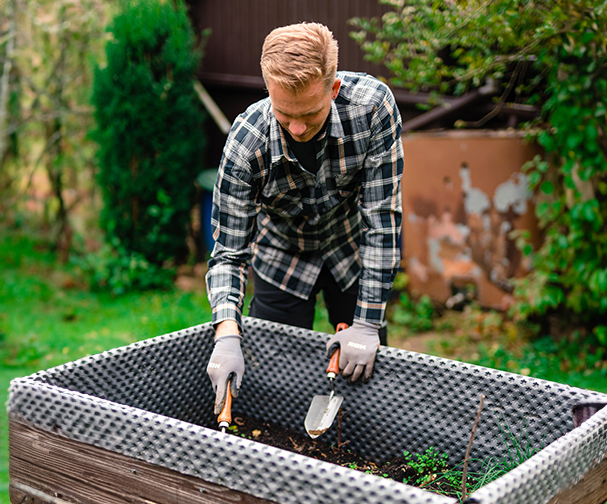 Mann mit kariertem Hemd und Gartenhandschuhen arbeitet mit Handwerkzeugen in einem Hochbeet im Garten; umgeben von Rasen und Struchern.