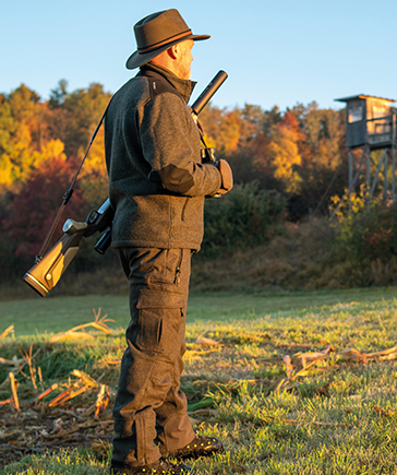 Une personne en tenue de chasse sombre avec chapeau se tient dans une prairie et porte un fusil � l’�paule, avec un mirador visible � l’arri�re-plan.